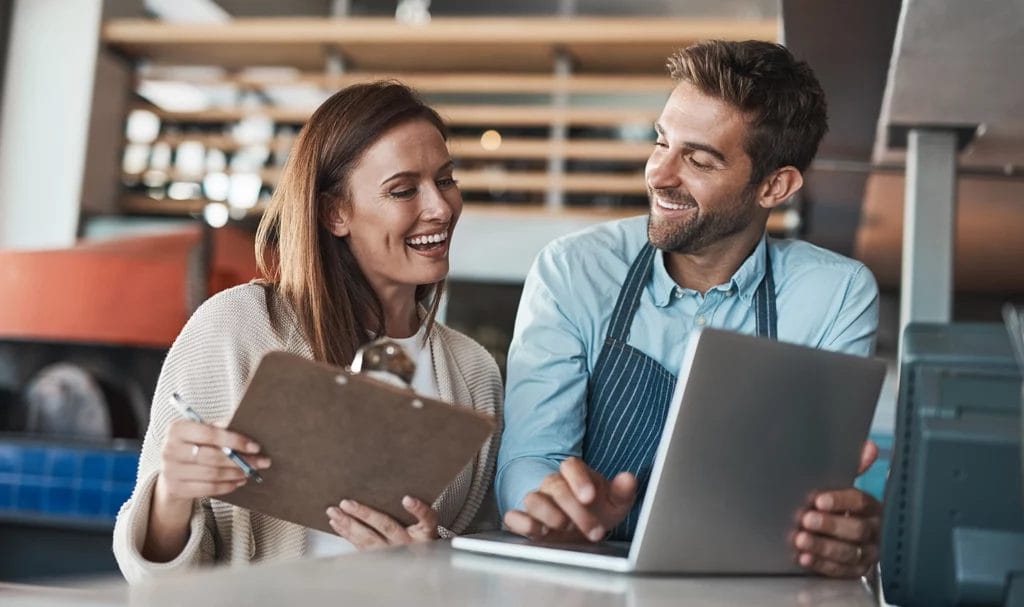 A small business consultant holding a clipboard happily smiles with their client as they go over information on a laptop.