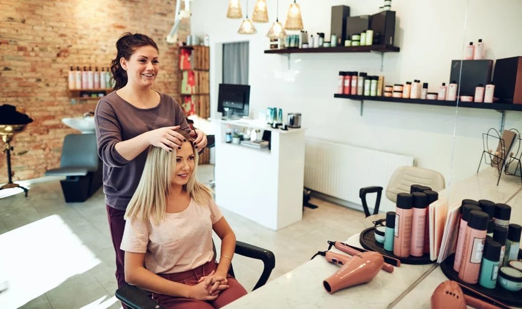 A hair stylist happily parts a client's hair while she sits in her salon chair at her station.