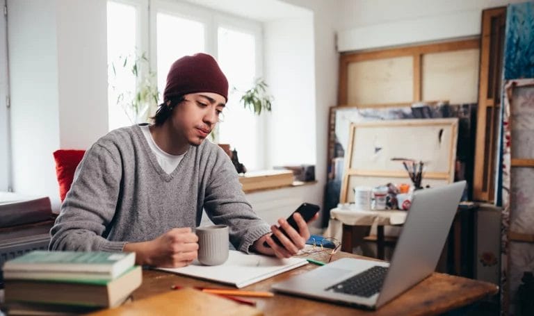 A small business owner is on his smartphone in his studio next to his laptop as he holds a mug of coffee.