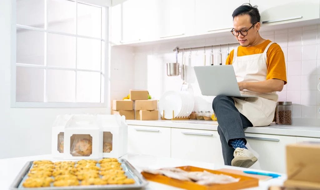 A home baker sits on the kitchen counter while using a laptop, across from an island full of pre-made and packaged baked goods.