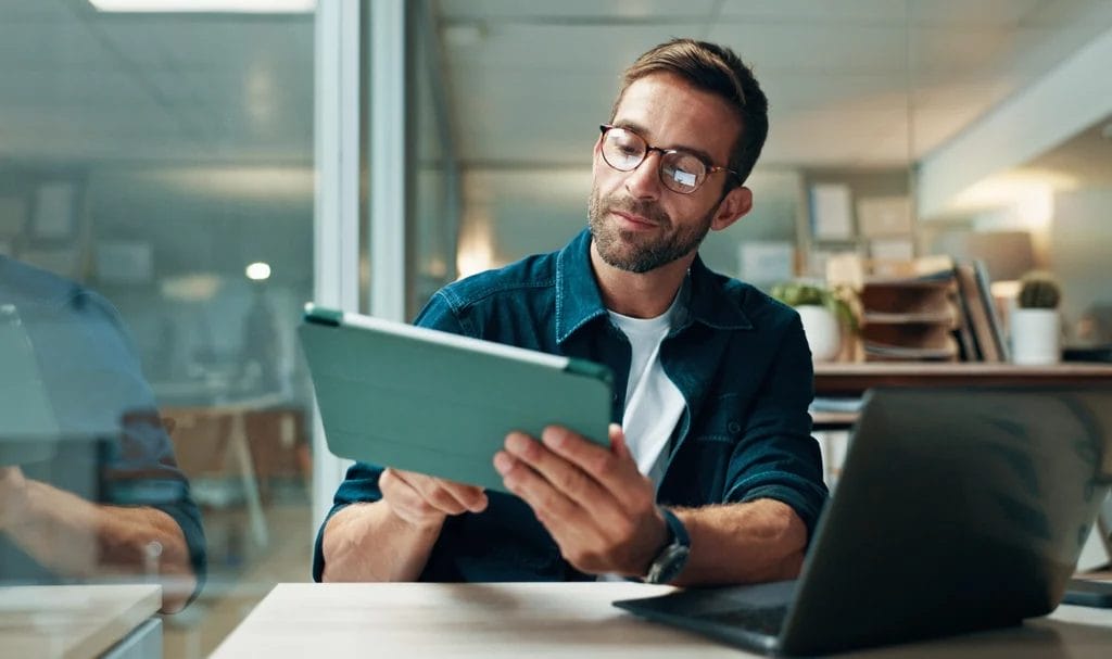 An entrepreneur is using a laptop with a teal case while working on business tasks on this device and a laptop.