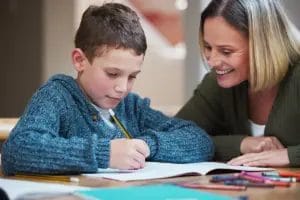 A woman with shoulder-length blonde hair helps a young male student in a blue sweater as he writes in a workbook at home.