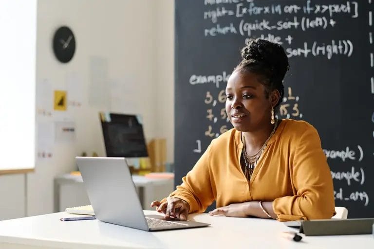 A woman in a yellow cardigan teaches online via a laptop with a chalkboard behind her.