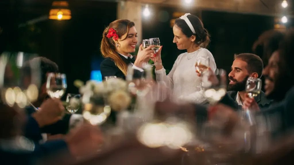 a big dinner party with two female hosts proposing a toast among guests and raising wine glasses over a dinner table