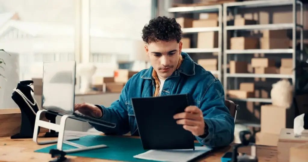 A small business owner wearing a denim jacket holds a black folder and types on a business laptop propped on a laptop stand while seated at a wooden desk in an inventory room.