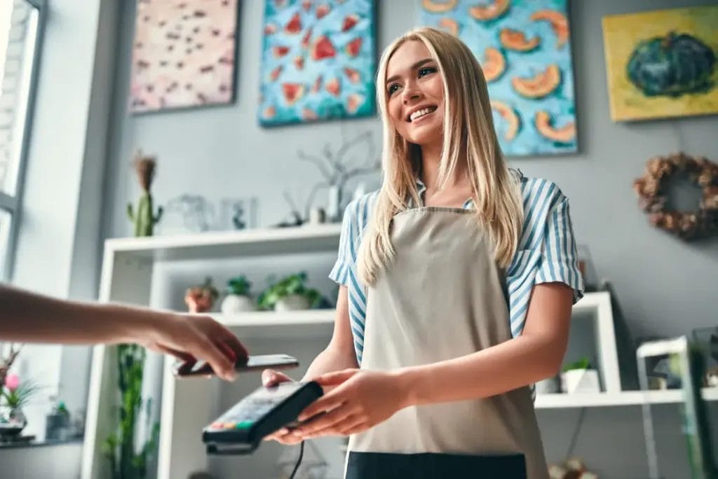 A small business owner wearing a teal striped top and a beige apron holds a card reader to accept payment from a customer in a boutique store with colorful artwork hanging on the wall behind her.