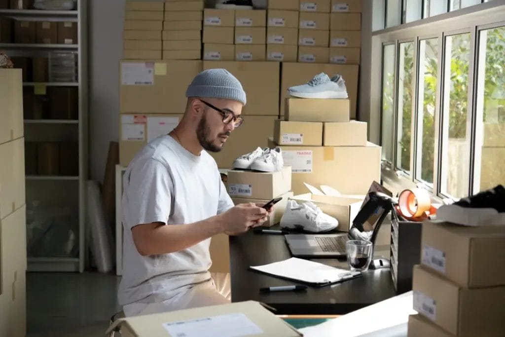 A small business owner wearing a gray beanie and a light gray t-shirt types on his mobile smartphone while seated at a desk with sneakers and shoeboxes stacked in an inventory room.