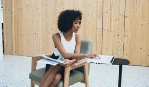 A small business owner wearing a white sleeveless blouse and a black skirt holds business papers and types on her laptop while seated at a green armchair in a modern office with wooden cabinets behind her.