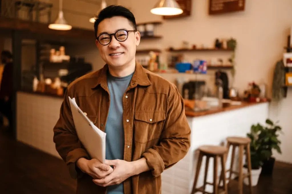 A business owner wearing round glasses and a brown jacket holds a contract in his arms and smiles in a modern cafe space.