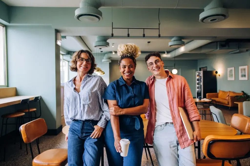 A group of business professionals smile and lean against a wooden table in a mint-colored shared office space.