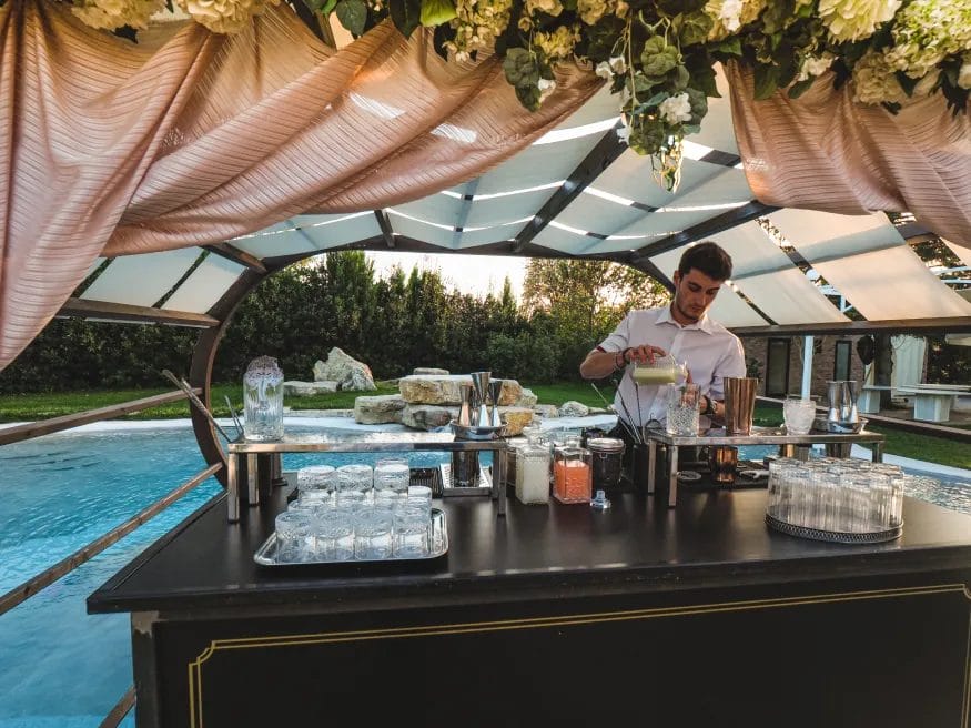A mobile bartender makes cocktails at a wedding bar stand under a pink canopy with white and green florals.