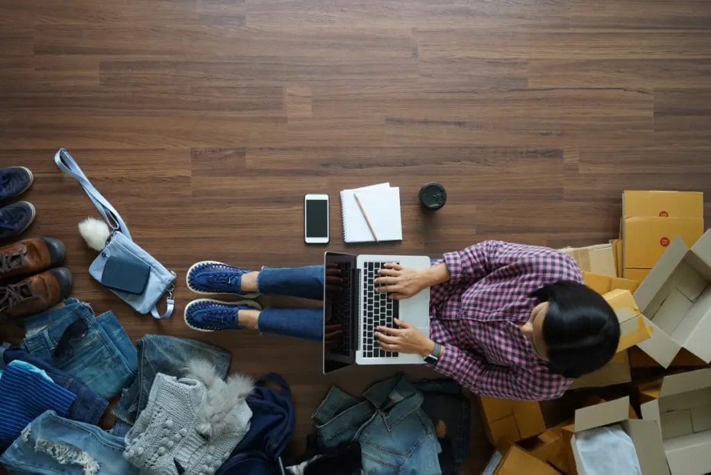 Top-down view of a small business owner working on a laptop while packaging clothes for shipping.