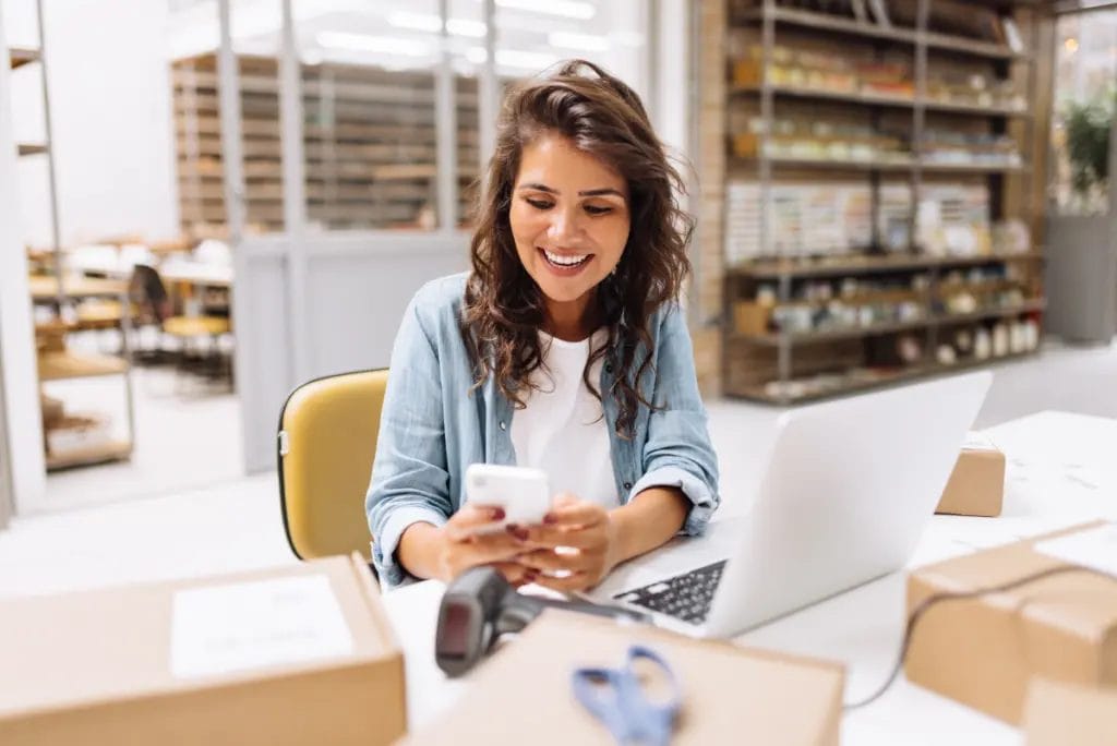 Business owner in brightly lit warehouse smiles down at her mobile phone while working.