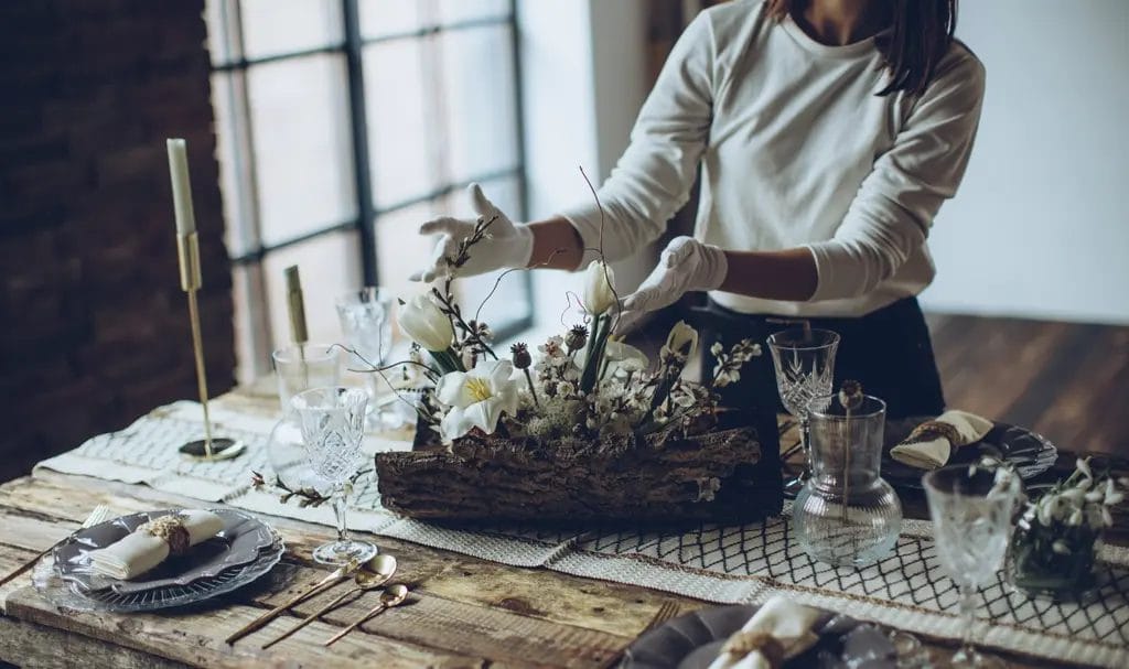 A close-up look at a party planner arranging a floral centerpiece on a set dinner table while wearing white gloves.