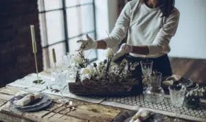 A close-up look at a party planner arranging a floral centerpiece on a set dinner table while wearing white gloves.