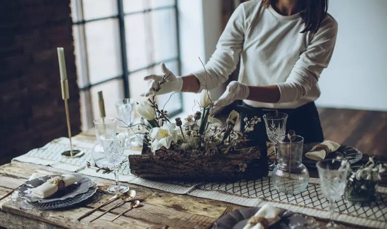 A close-up look at a party planner arranging a floral centerpiece on a set dinner table while wearing white gloves.