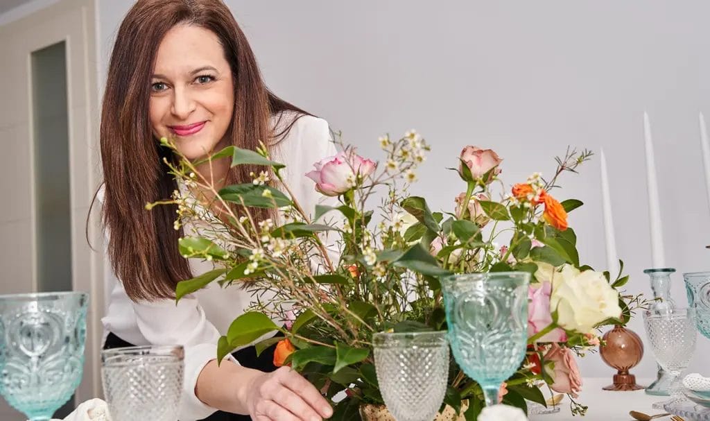 An event planner smiles at the camera as she arranges a floral centerpiece on a table set for a nice dinner.