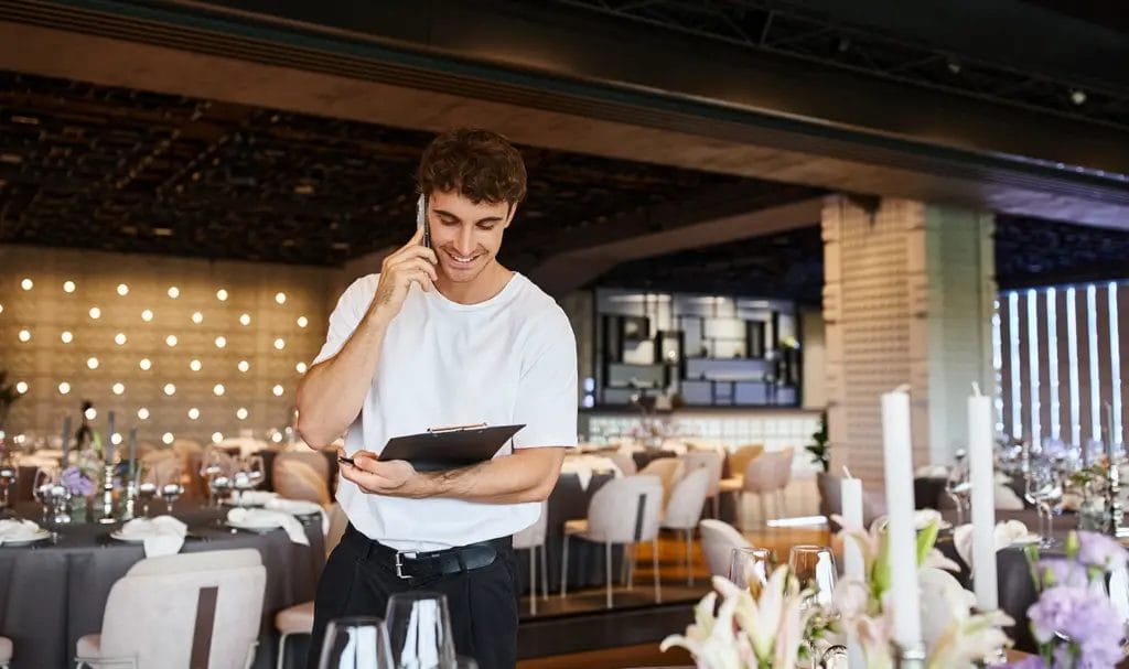 A party planner happily speaks on the phone as he looks down at a clipboard and walks through a decorated venue before an event.
