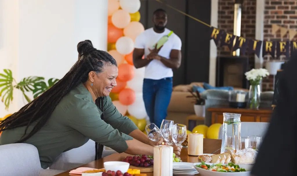 A party planner joyfully sets a handful of wine glasses on a set table for a retirement party in a guest's home.
