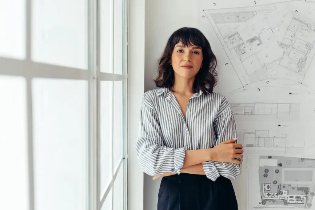 A small business owner wearing a striped shirt and blank slacks folds her arms and leans against an office wall next to a paned window.