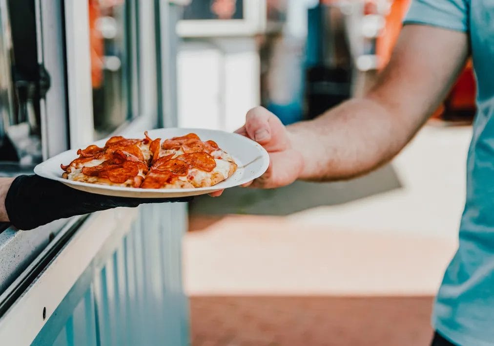 A food truck owner hands a mini pepperoni pizza on a paper plate to a customer through the food truck window.