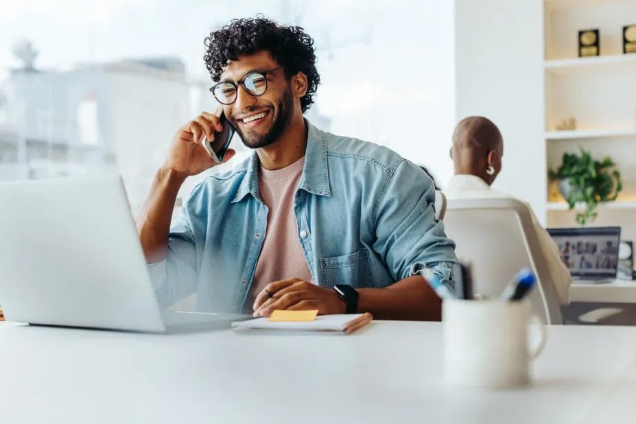A small business owner wearing a denim shirt and glasses talks into his smartphone while sitting at a desk with his laptop and notepad in front of him.