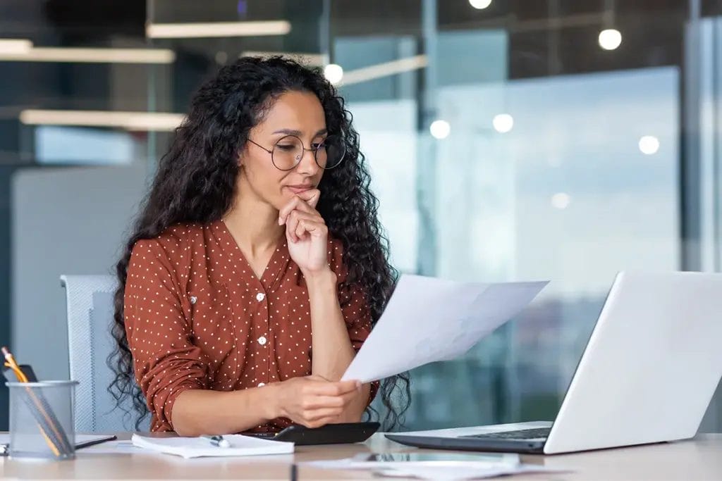 A professional woman in a brown button-up, polka dot shirt reads a document while working on her laptop in a conference room.