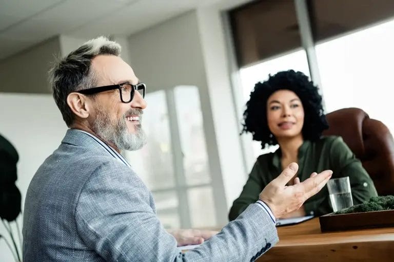 An older man with a beard and glasses wearing a gray blazer smiles as he talks to coworkers in a corporate office setting.