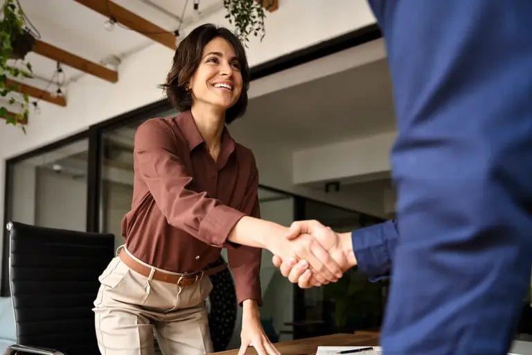 A woman wearing a brown button-down shirt and slacks rises from her seat as she shakes hands with another employee wearing a blue shirt in a corporate office setting.