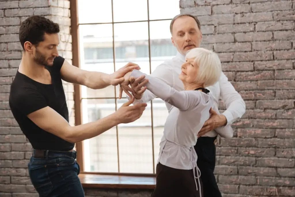 A ballroom dance teacher guides the form of a ballroom dance couple in a dance studio with brick walls and a paned glass window.