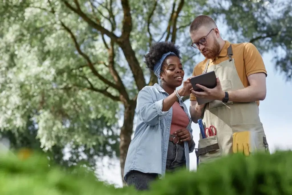 Two landscaping professionals discuss a design project while looking at a business tablet outdoors with a large tree behind them.
