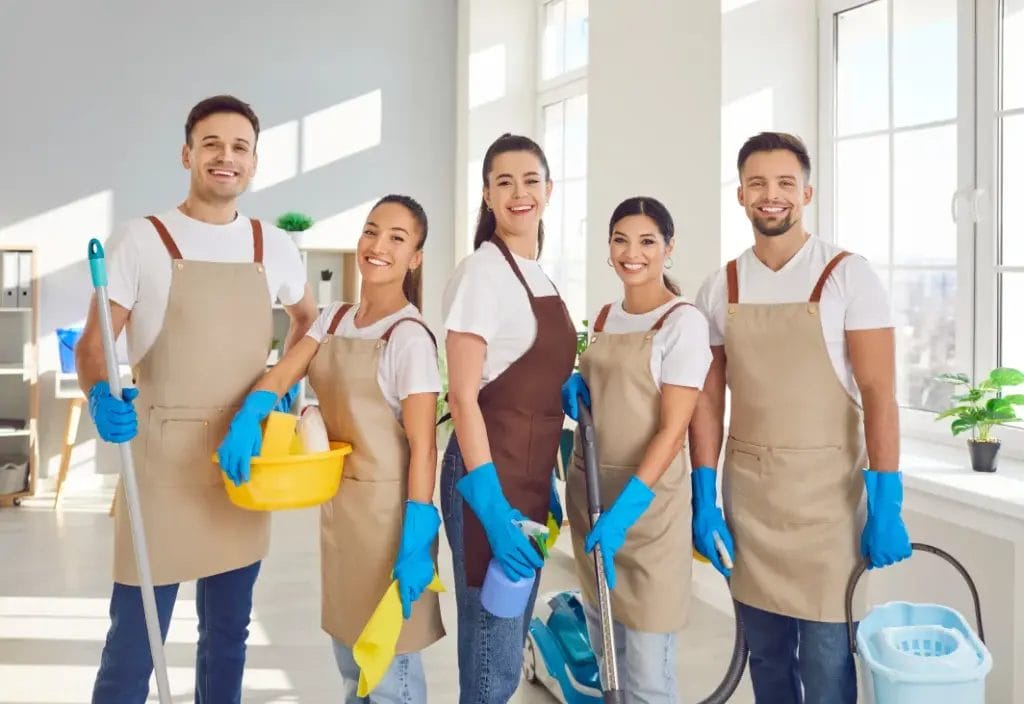 A group of cleaning professionals wearing white t-shirts, beige aprons, and blue gloves hold cleaning supplies and smile while standing in a large room with glass windows.