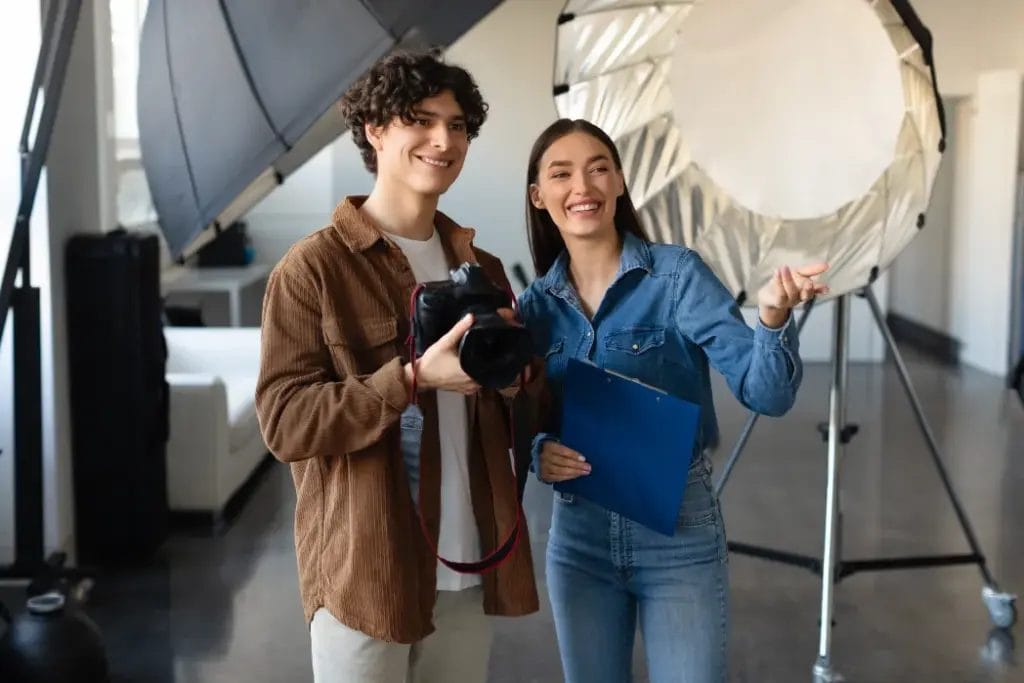 A photographer wearing a brown shirt and holding a camera smiles and speaks with another photographer wearing a denim outfit on a photography set with modifier lighting equipment in the background.