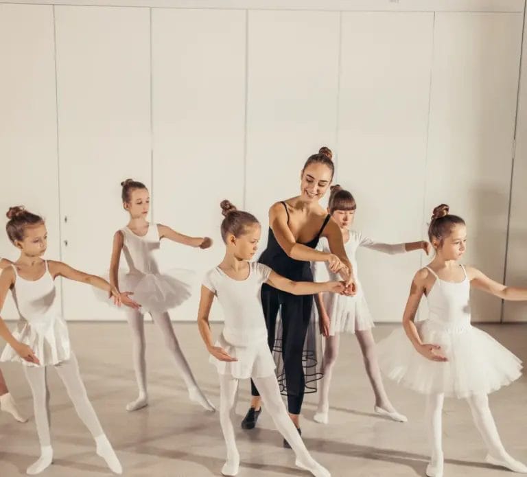 A ballet dance teacher guiding her students' third-position arms during a dance class in a bright, white studio.