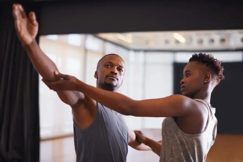 A dance teacher wearing a gray tank top guides the arms of a dance student during a dance class in a studio with mirrors and black curtains in the background.