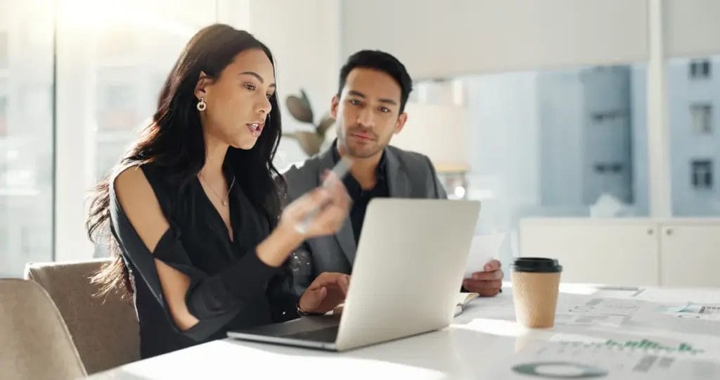 A business consultant wearing a black top gestures to a silver-toned laptop while speaking with a client as they sit at a table with papers in a modern office setting.