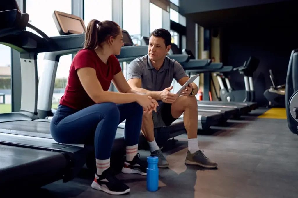 A personal trainer wearing a gray polo shirt and shorts holds a tablet and speaks with a client wearing a red shirt and blue leggings while they sit on treadmills in a gym.