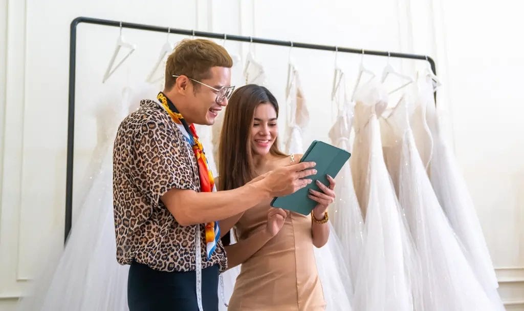 A wedding planner reviews details on a tablet with a wedding dress designer inside a dress showroom.