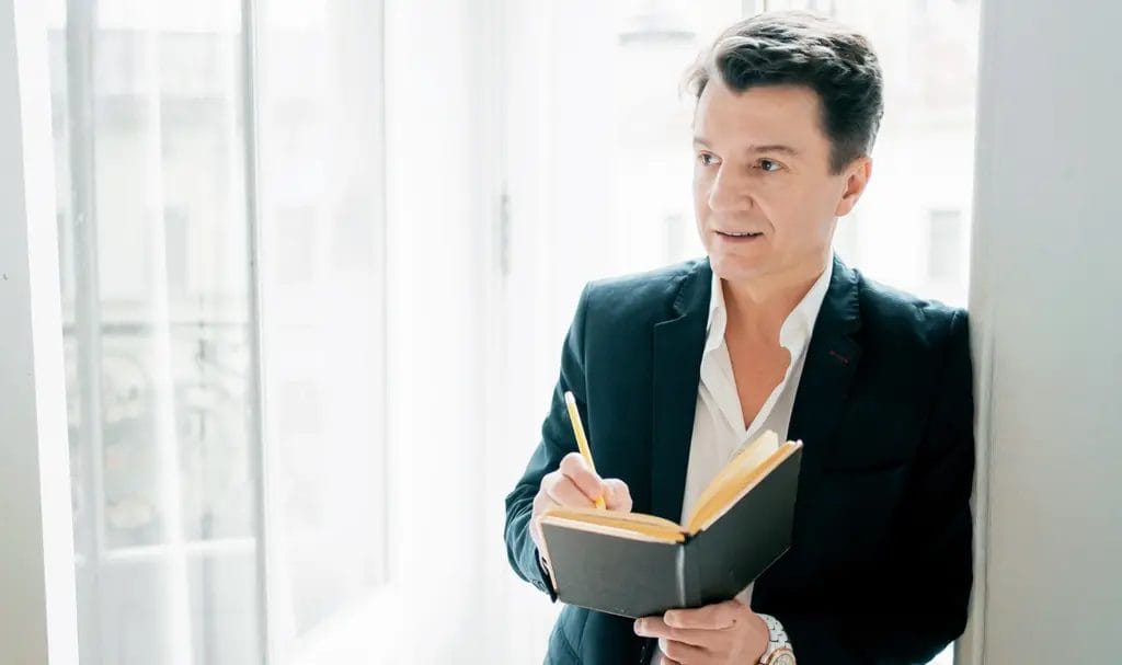 An event planner leans on the doorframe of a well-lit event room as he takes notes in a notebook and observes the space for a potential upcoming event.