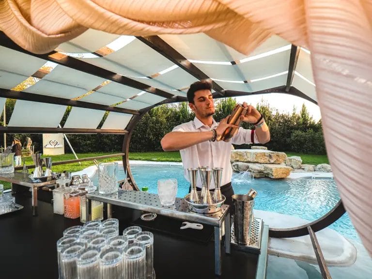 A male bartender shakes a cocktail shaker at a pop-up bar in front of a water feature at a wedding.