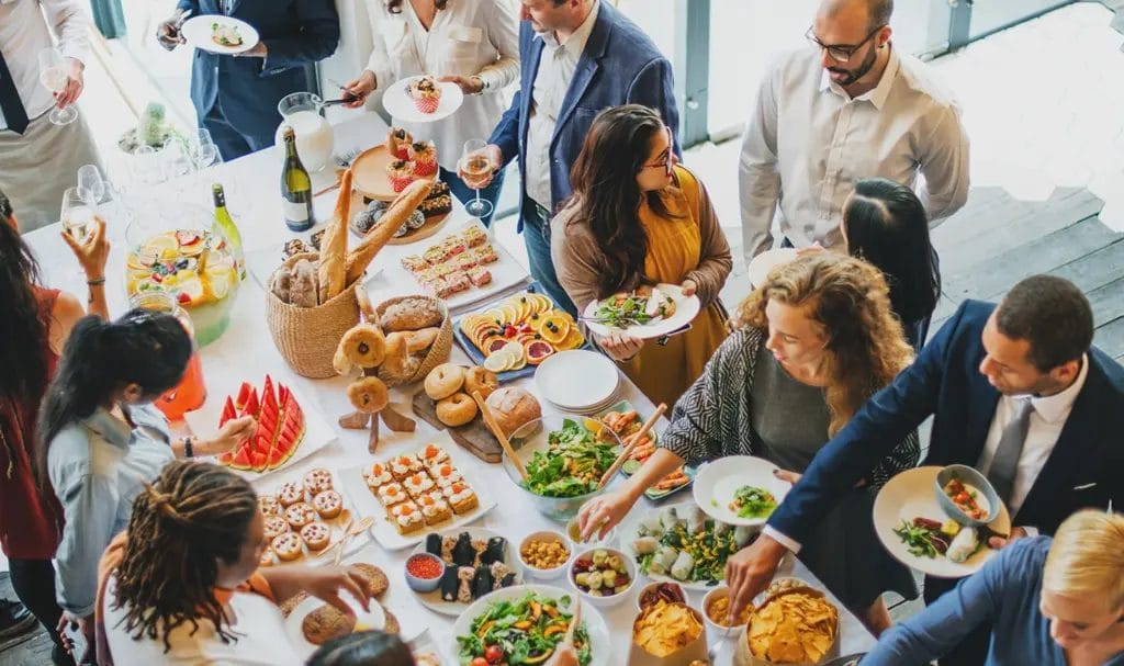 An overhead view of a corporate gathering where employees fill plates around a catered meal table.