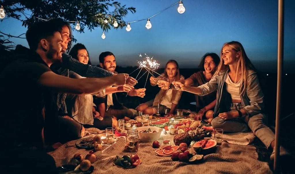 A group of friends with sparklers gather around a blanket full of food as they celebrate a private event.