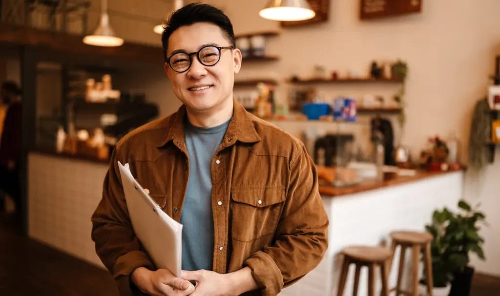 A happy business owner smiles for the camera inside a warmly lit cafe while holding paperwork in his arms.