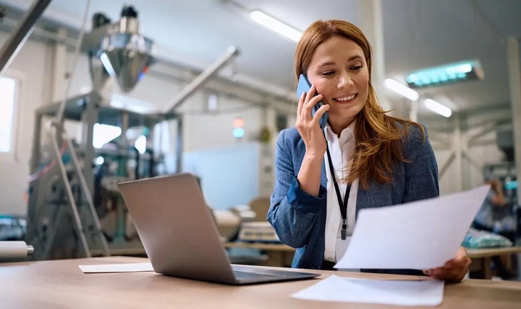 A cheerful businesswoman is in an industrial manufacturing office space where she is sitting at a desk with a laptop and paperwork as she happily speaks on a smartphone.