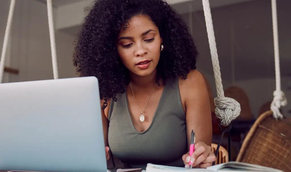 A businesswoman carefully reviews information in a notebook with information on her laptop.