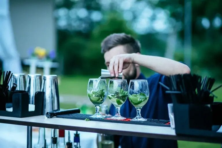 A male bartender pours a shot of liquor into a cocktail next to two others on his bar mat at an outdoor event.