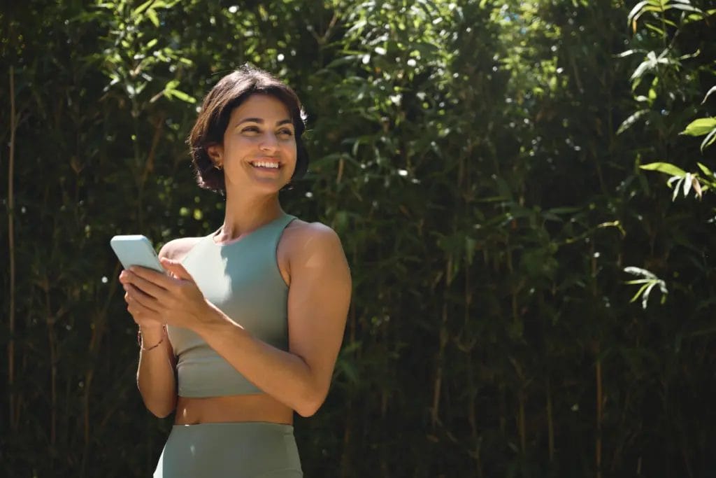 smiling yoga and fitness instructor in light green athletic wear standing outdoors in the sun, in front of a wall of out-of-focus trees.