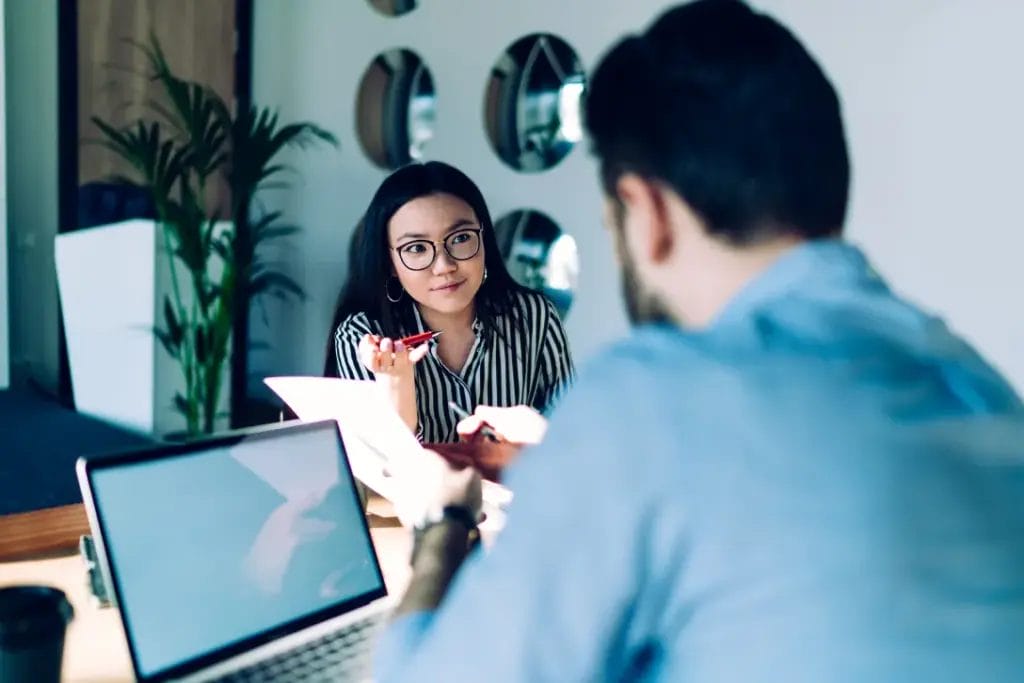 A marketing consultant speaks with a client as they sit a table in an office with a green plant and multiple round mirrors in the background.