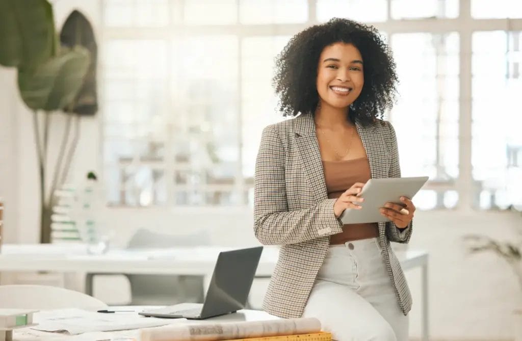 A professional consultant wearing a houndstooth blazer and white pants smiles while holding a business tablet and leaning against a desk in a bright modern office.