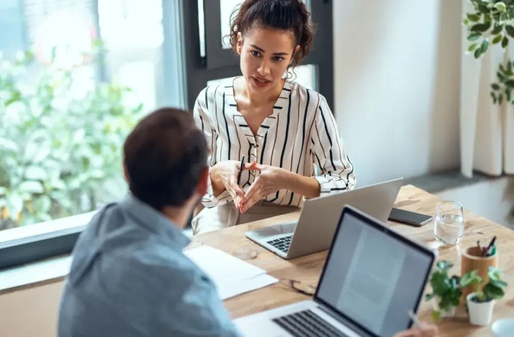 A business consultant wearing a white and navy striped blouse speaks with a client as they meet at a wooden desk in a bright office with a large window overlooking a tree in the background.